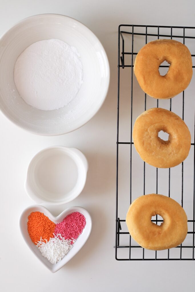 Deep fried doughnuts resting on baking rack with bowl of sprinkles on the side