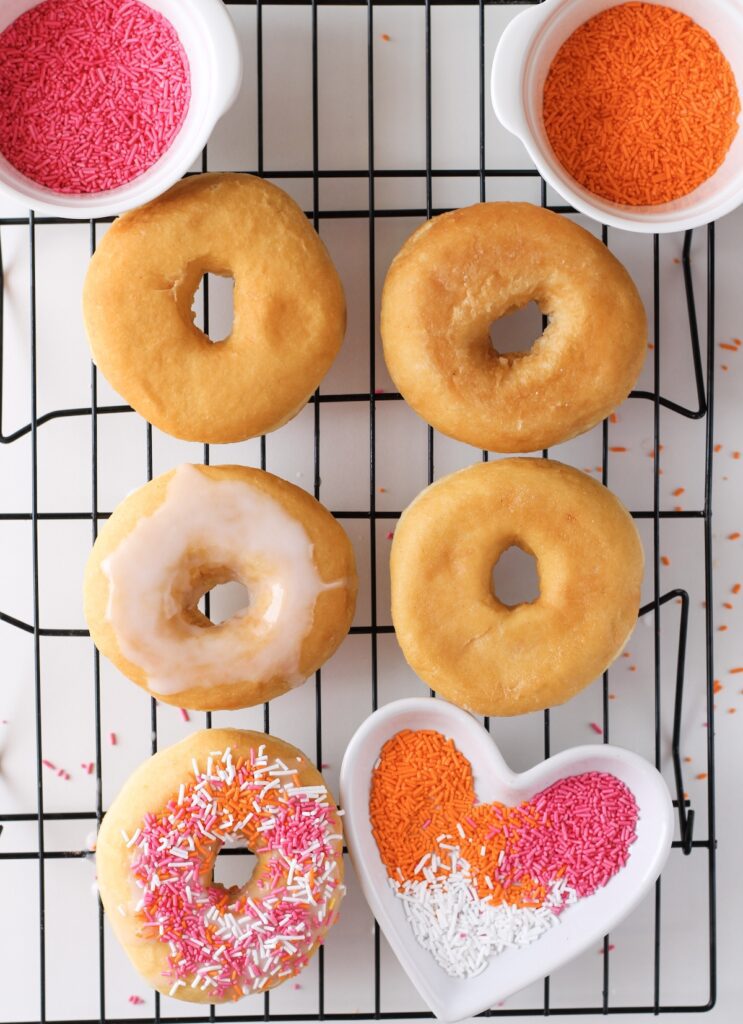 Donuts resting on baking rack some pieces with icing and sprinkles on top with bowl of sprinkles on the side
