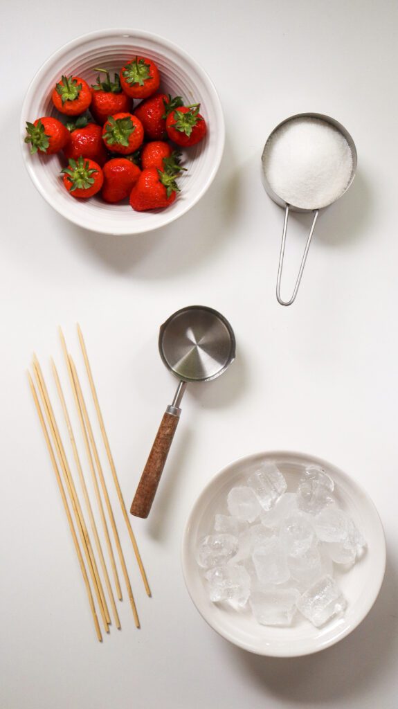 Fresh strawberries inside a big bowl, a bowl of ice, measuring cup, sugar, and barbeque sticks