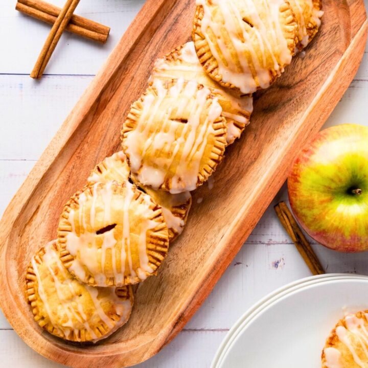 A wooden tray filled with round mini apple hand pies drizzled with white icing, surrounded by fresh apples and cinnamon sticks on a white rustic table