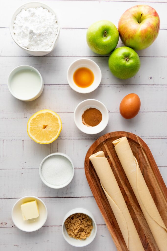 Flat lay of ingredients for mini apple hand pies including green and red apples, pie dough rolls, powdered sugar, butter, brown sugar, granulated sugar, cinnamon, vanilla, lemon, milk, and an egg