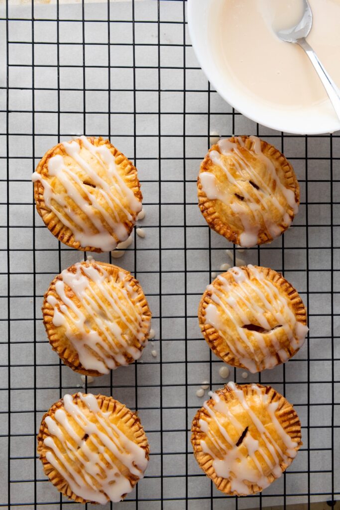 Six mini apple hand pies on a wire rack, generously drizzled with icing and a bowl of glaze visible in the corner