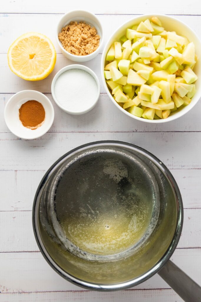 Ingredients like chopped apples, lemon, sugars, and cinnamon placed around a metal saucepan with melted butter