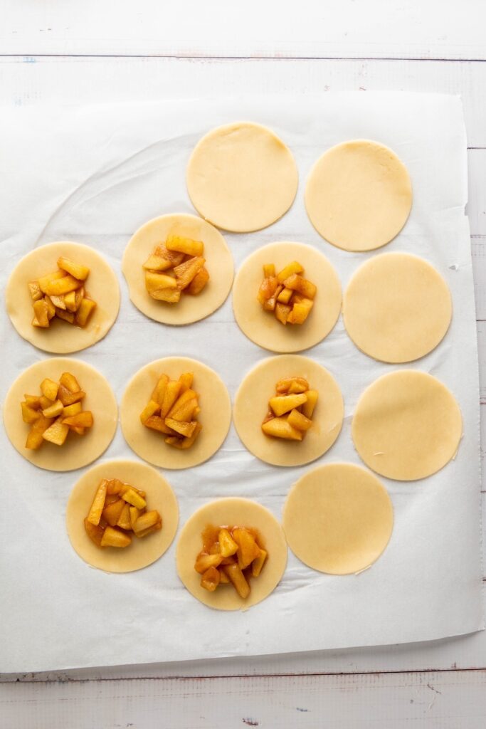 Round pie dough cutouts filled with cinnamon apple mixture, spaced on parchment paper and ready to be sealed