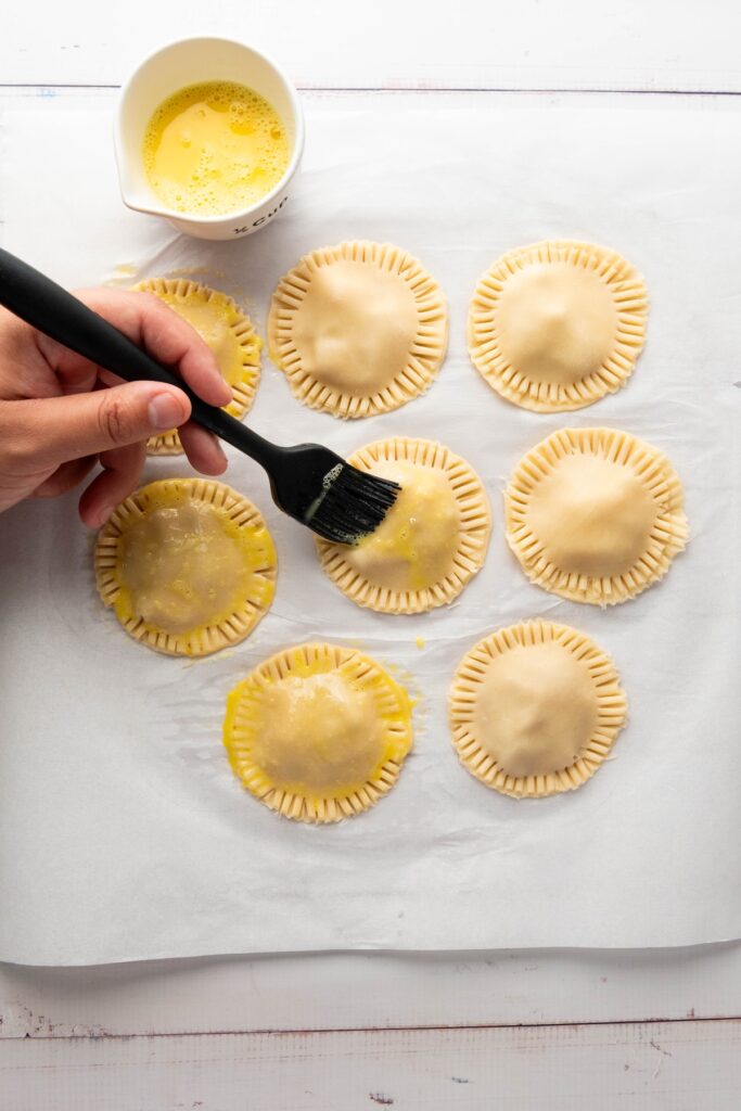 Hand brushing an egg wash onto the sealed apple hand pies, which have crimped edges and sit on parchment