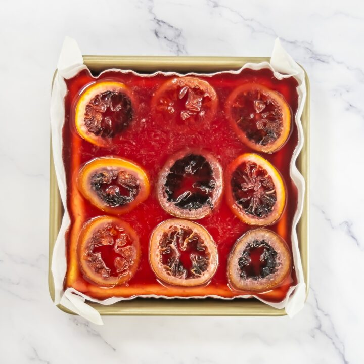A full view of the decorated cheesecake bars in a square pan with evenly placed candied blood orange slices on top of vibrant red jelly