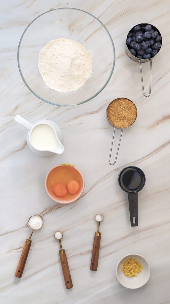 Overhead shot of ingredients for waffles featuring flour, brown sugar, blueberries, milk, eggs, lemon zest, oil, and small spoons of baking powder, baking soda, and salt.