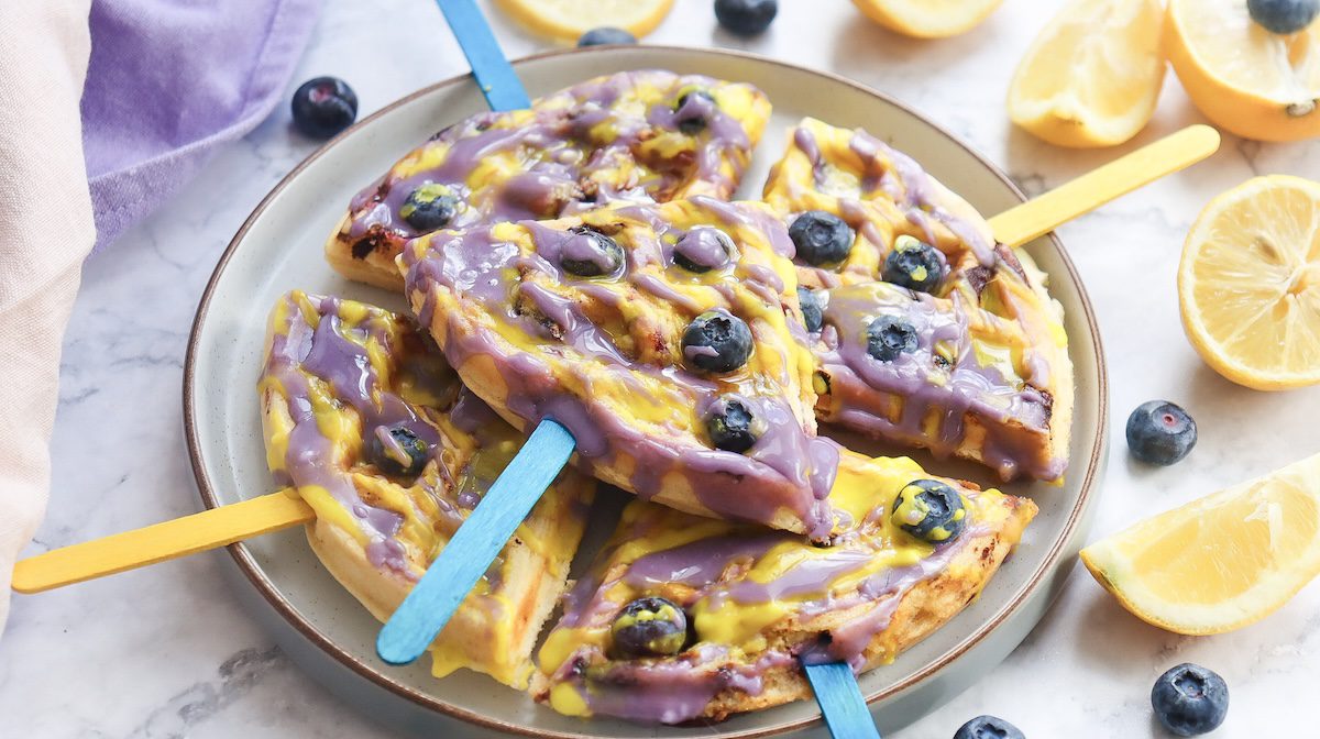 A plate of blueberry lemon waffle pops, each waffle slice on a colorful stick and drizzled with purple and yellow icing, garnished with whole blueberries and surrounded by lemon wedges
