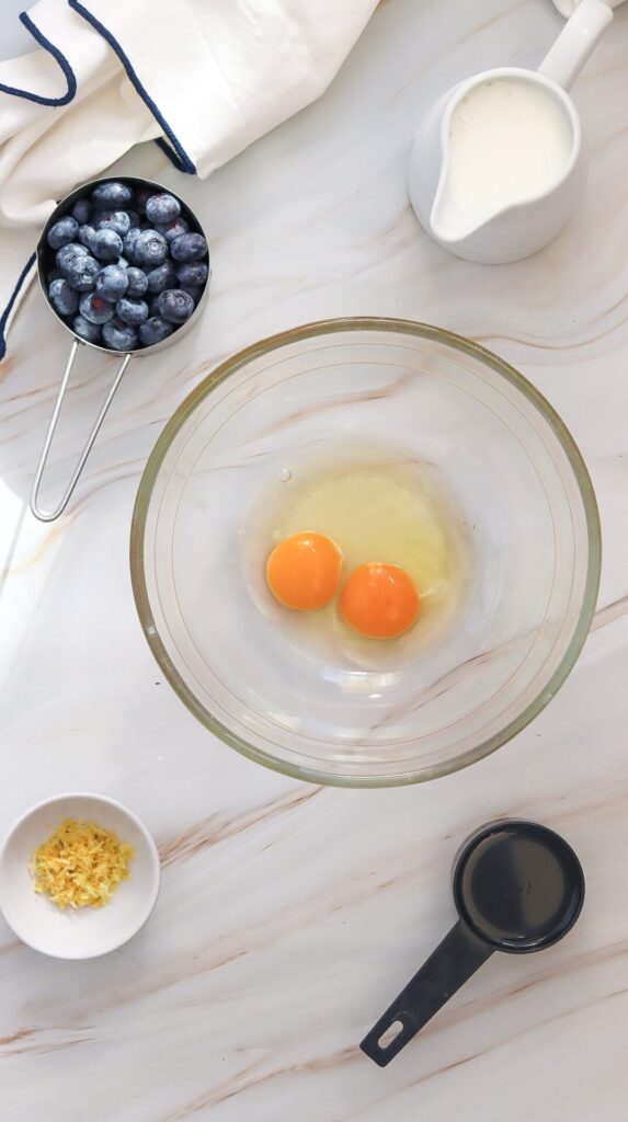 A bowl containing two cracked eggs surrounded by other wet ingredients and mix-ins like milk, oil, lemon zest, and blueberries