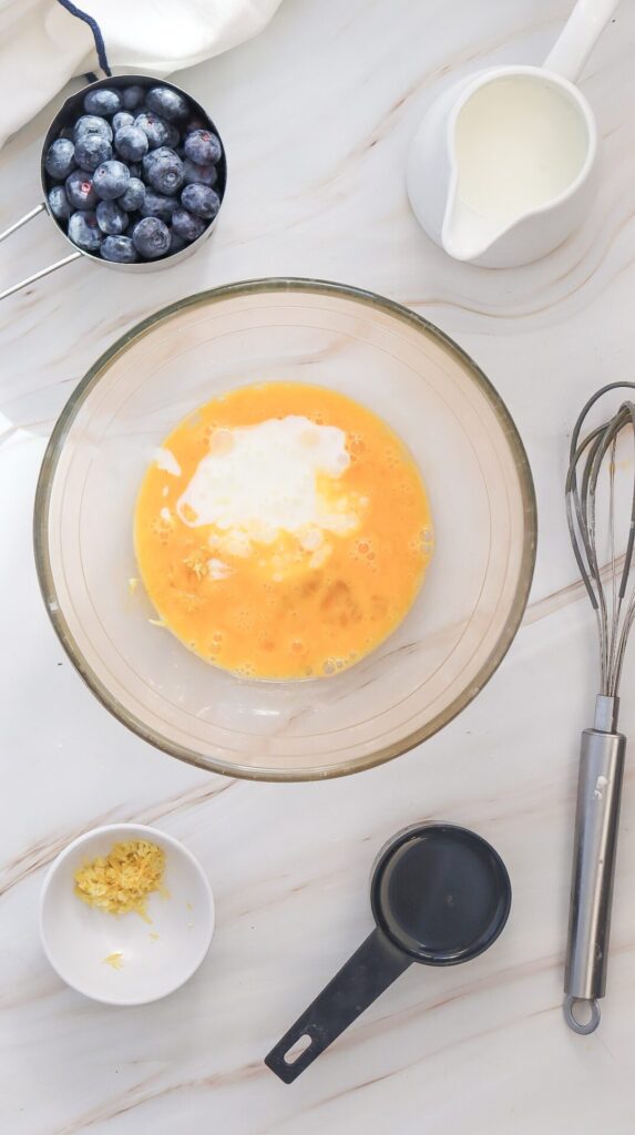Beaten egg mixture in a bowl with added milk and lemon zest, with other ingredients and a whisk nearby, mid-recipe prep
