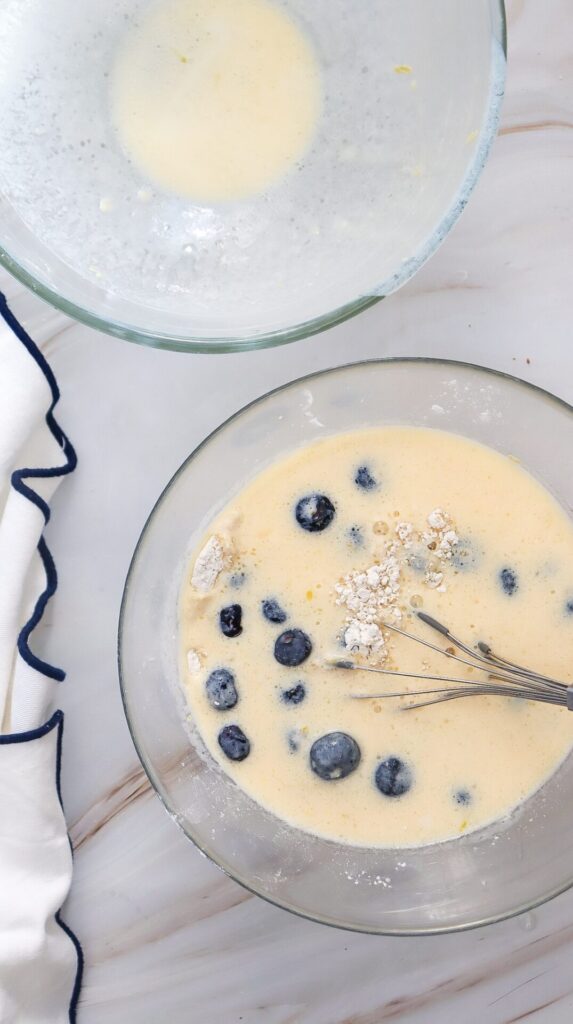 Mixing bowl filled with waffle batter that includes whole blueberries and bits of flour not yet fully combined, with a whisk partially in frame