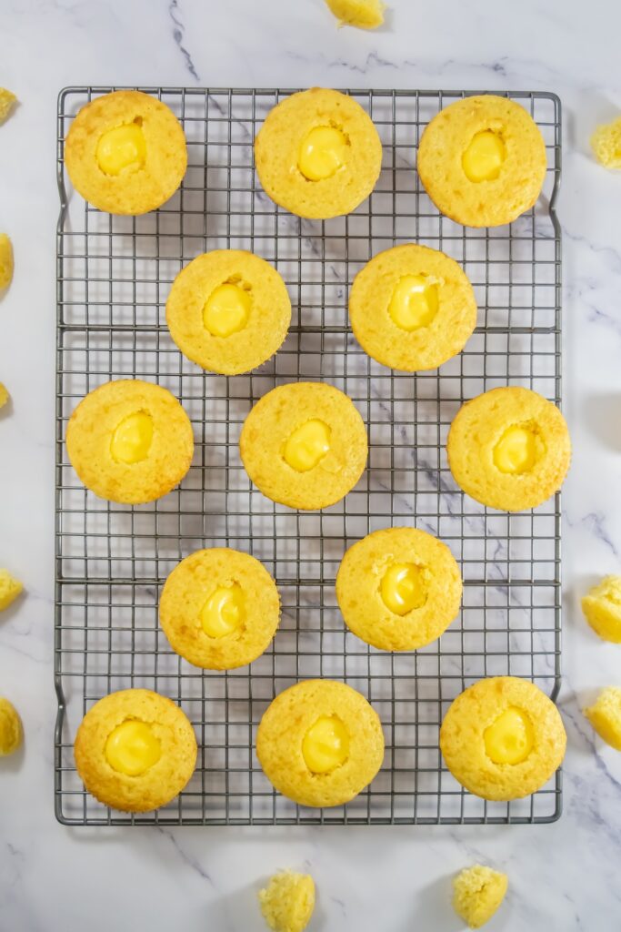 Top-down view of freshly baked yellow cupcakes on a cooling rack, each filled with creamy yellow custard in the center, surrounded by small chunks of removed cake tops