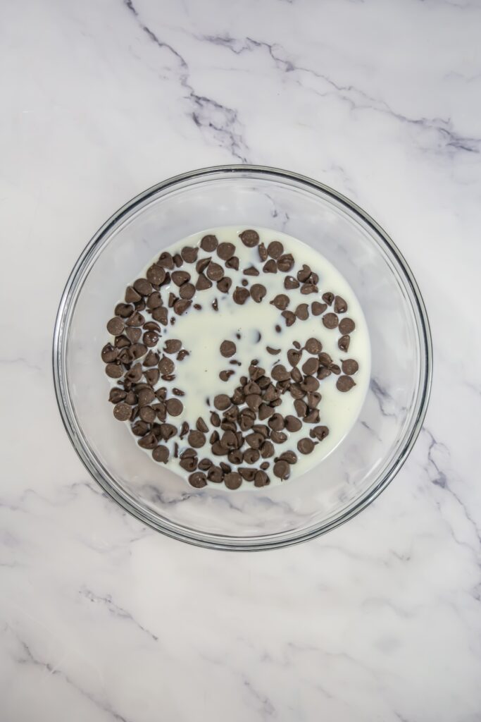 A clear bowl filled with chocolate chips and milk before melting, prepared for ganache topping