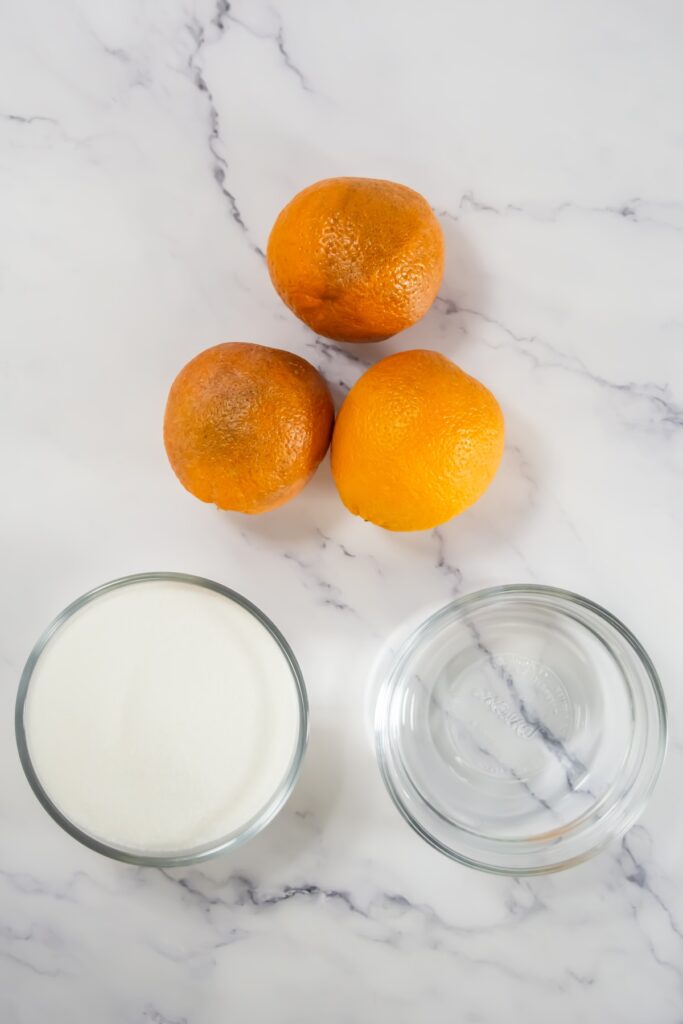 Three whole blood oranges rest on a marble surface beside two glass bowls—one filled with granulated sugar and the other with water, the basic ingredients for making candied oranges