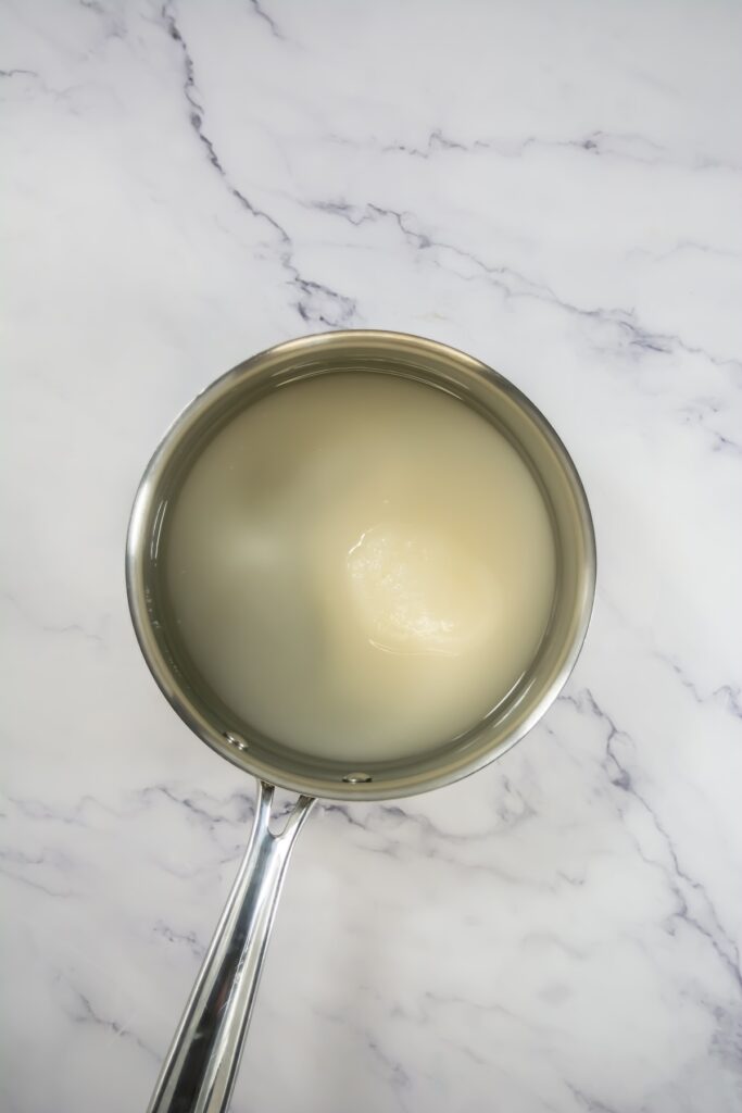 A saucepan filled with a pale sugar and water mixture on a marble countertop, showing the beginning stage of the simple syrup used to candy the oranges