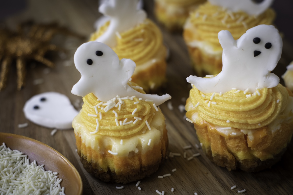 Mini cheesecakes layered in white, orange, and yellow to resemble candy corn, topped with orange frosting, white sprinkles, and fondant ghosts with cute black eyes