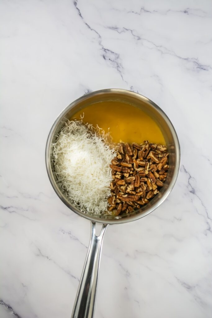 Overhead view of shredded coconut and chopped pecans added to the golden frosting base in a saucepan, ready to be mixed for traditional German chocolate cupcake topping