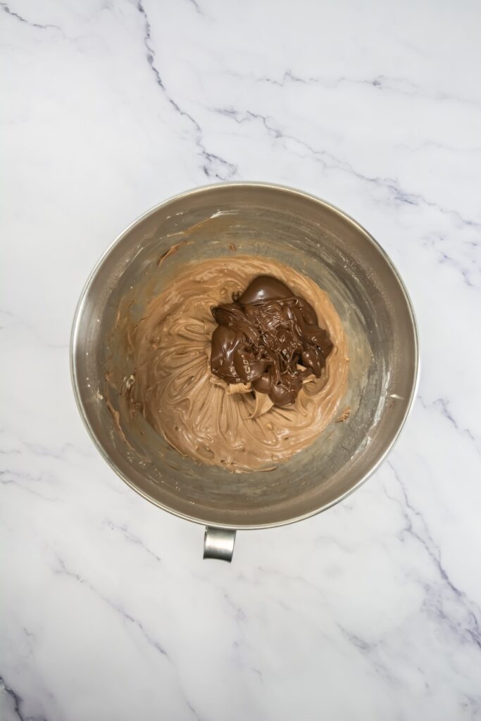 A mixing bowl of chocolate frosting with a generous dollop of melted chocolate added on top, ready to be blended in