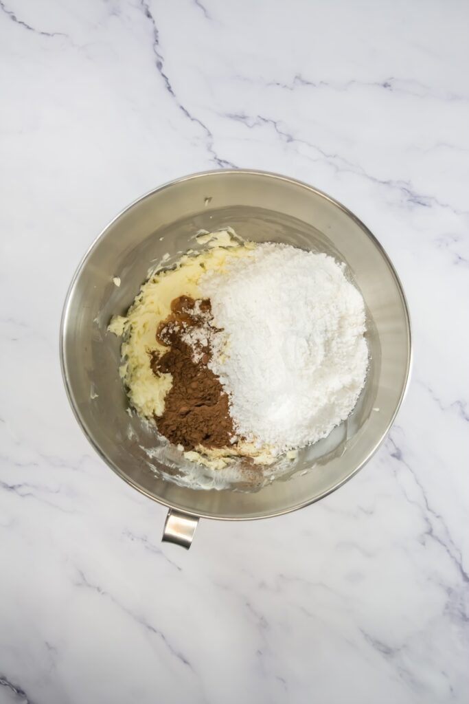 Mixing bowl showing cocoa powder and powdered sugar piled over the creamed butter and cream cheese, about to be mixed for chocolate frosting