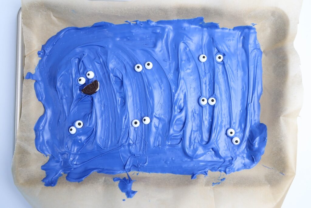 A baking tray lined with parchment paper holding a thick layer of blue candy coating decorated with scattered pairs of candy eyes and one sandwich cookie mouth, resembling playful Cookie Monster faces starting to take shape
