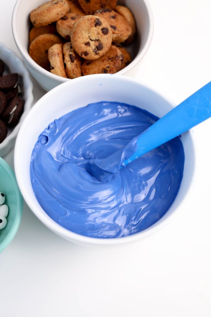 Close-up of blue melted candy in a bowl with a blue spatula, surrounded by bowls of mini cookies and candy eyes, showing preparation for decorating