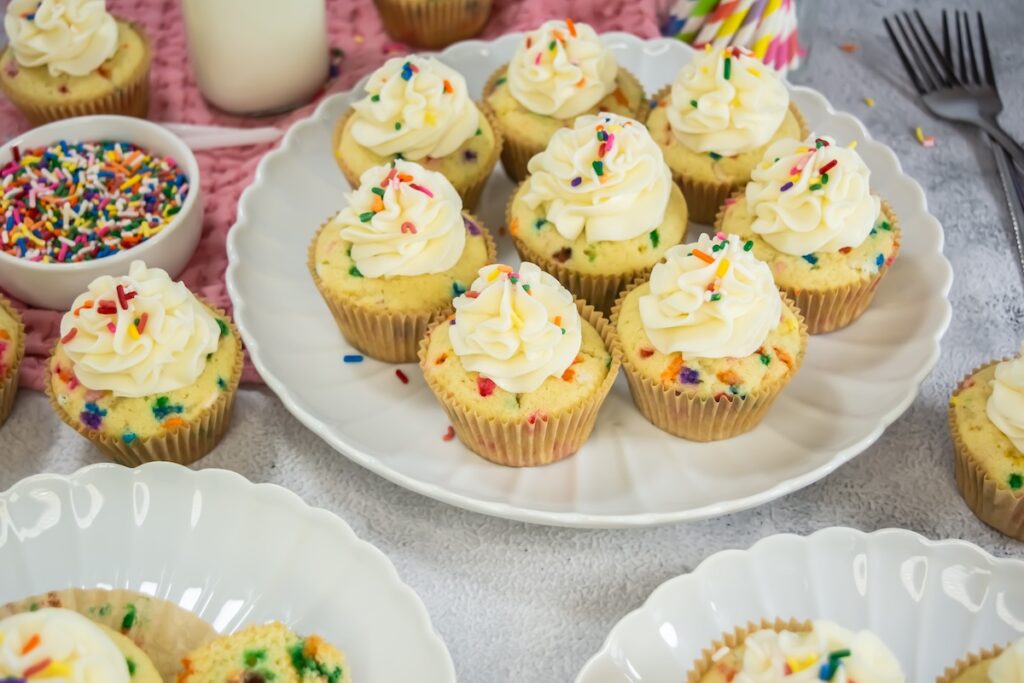 Overhead view of a platter filled with frosted funfetti cupcakes topped with rainbow sprinkles, surrounded by forks, a bowl of extra sprinkles, and colorful straws on a gray textured surface