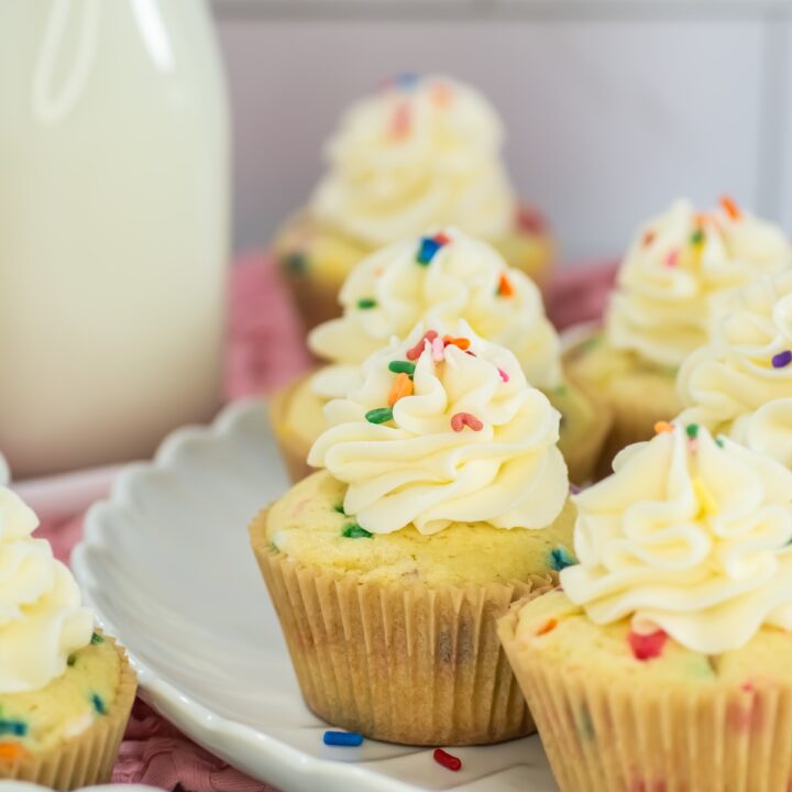Close-up of frosted funfetti cupcakes topped with swirled vanilla buttercream and colorful sprinkles, arranged on a white plate with a bottle of milk in the background