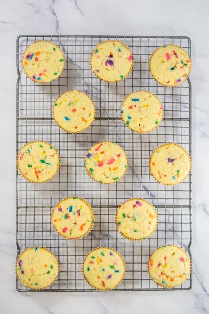 Baked funfetti cupcakes cooling on a wire rack, with bright specks of rainbow sprinkles visible in the golden tops