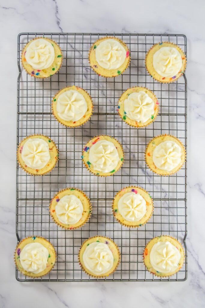 Overhead view of frosted funfetti cupcakes cooling on a wire rack, with white swirled buttercream piped on top of each colorful sprinkle-filled cupcake