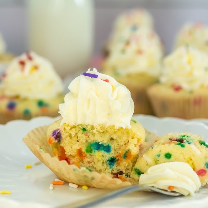Close-up of a funfetti cupcake with colorful sprinkles, topped with a swirl of vanilla frosting and partially eaten to reveal the vibrant sprinkles inside
