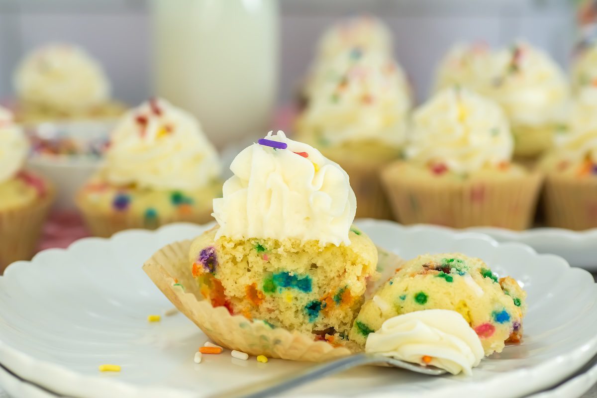 Close-up of a funfetti cupcake with colorful sprinkles, topped with a swirl of vanilla frosting and partially eaten to reveal the vibrant sprinkles inside