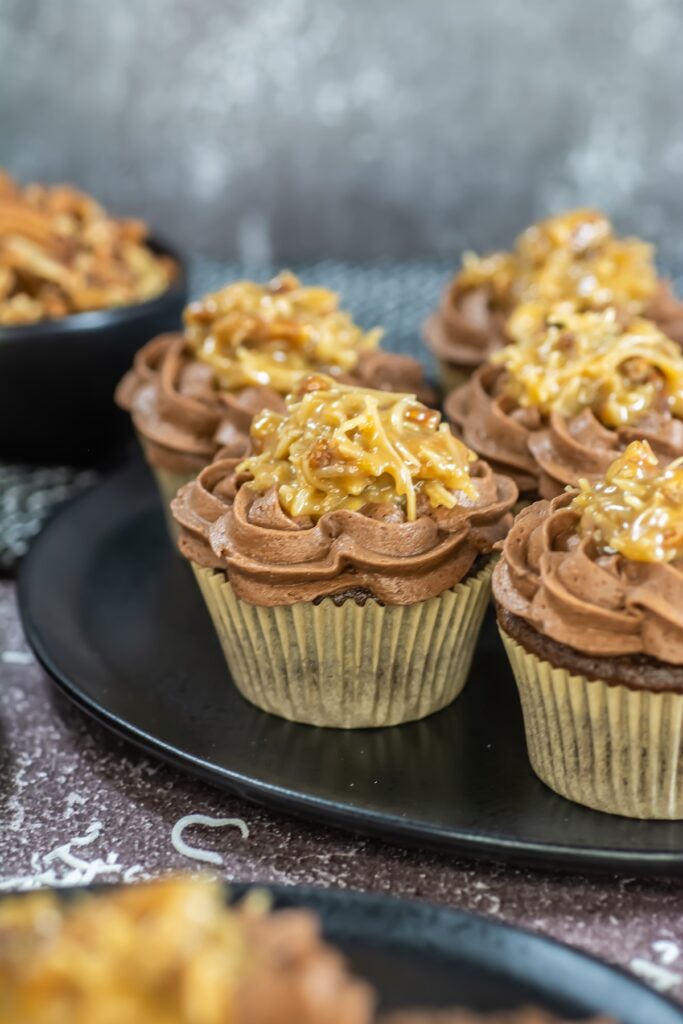 Close-up of frosted German chocolate cupcakes topped with a swirl of chocolate buttercream and a spoonful of coconut-pecan filling on black plates