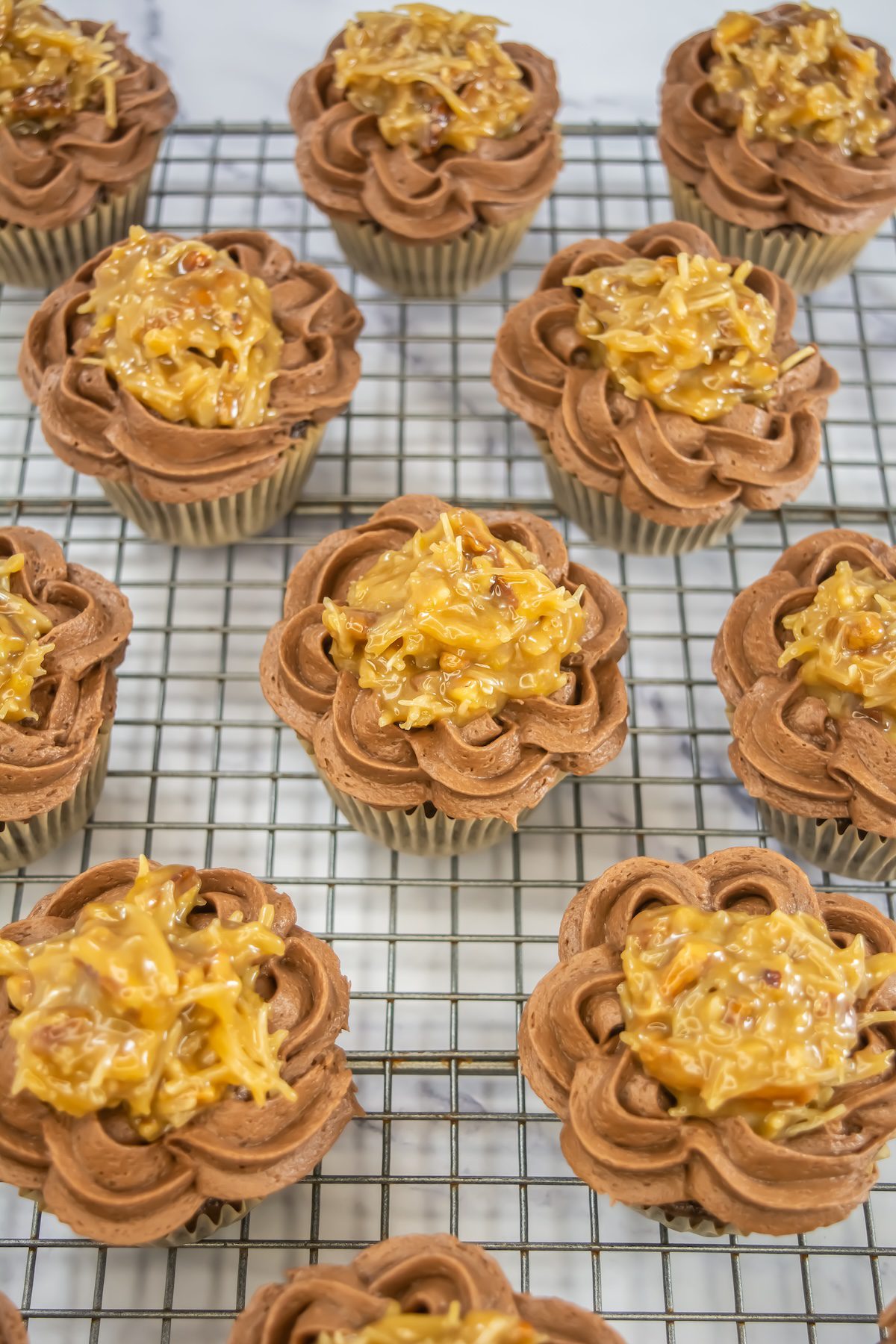 Overhead view of multiple German chocolate cupcakes on a wire rack, each decorated with piped chocolate frosting and coconut-pecan topping