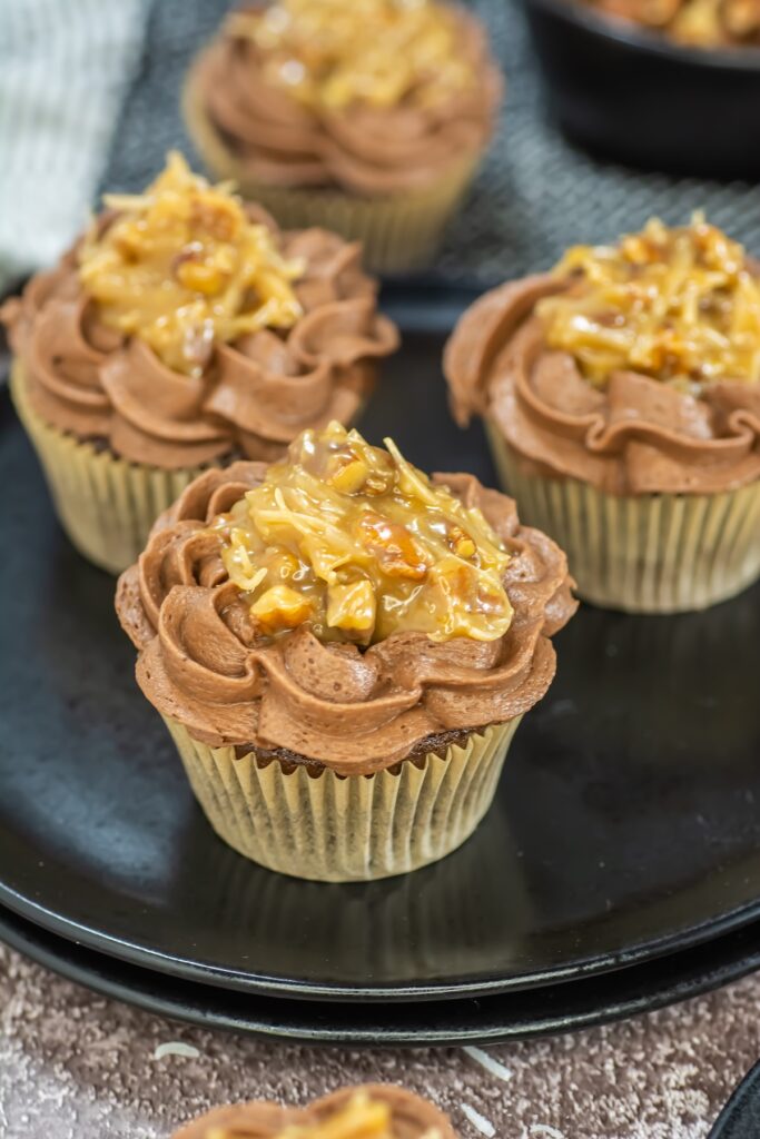 Close-up angled view of German chocolate cupcakes on a black plate, featuring rich chocolate frosting and a golden coconut-pecan topping
