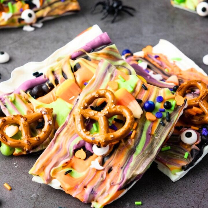 Close-up of Halloween candy bark pieces on a dark surface, featuring swirls of orange, green, purple, and black chocolate, topped with pretzels, candy corn, and festive sprinkles