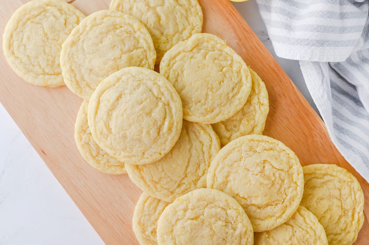 A batch of freshly baked lemon sugar cookies arranged on a wooden board next to a striped dish towel