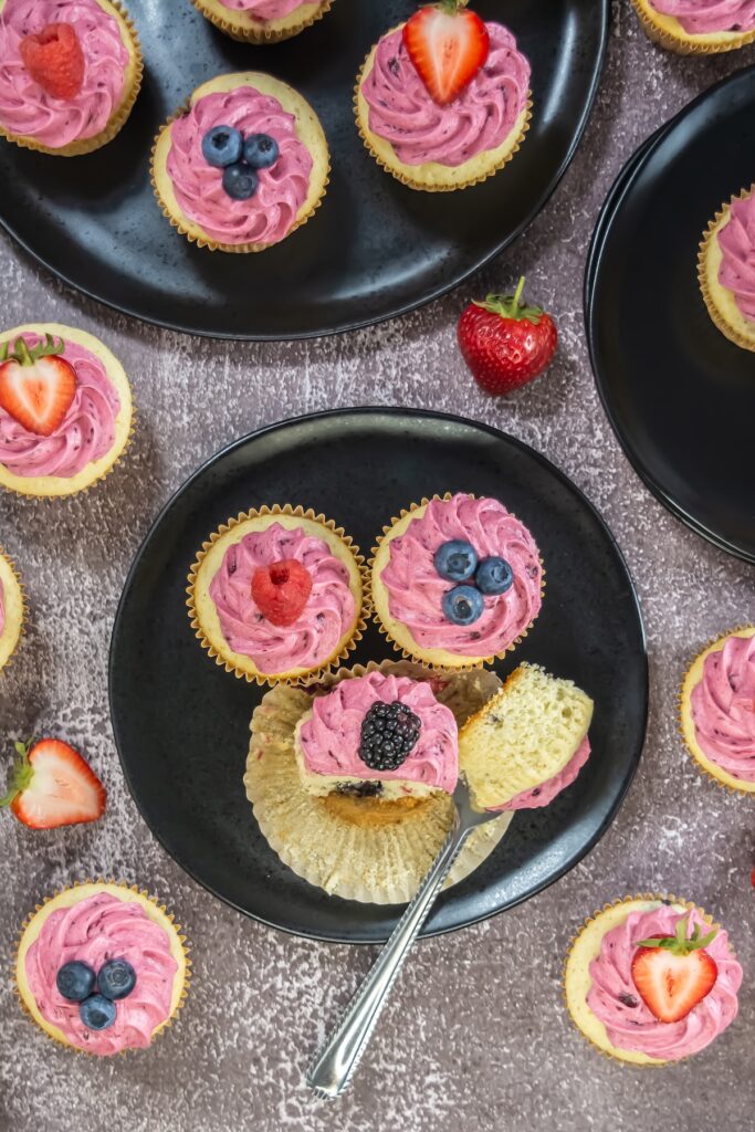 Overhead view of assorted mixed berry cupcakes topped with pink berry buttercream and fresh fruit, arranged on dark plates against a textured background