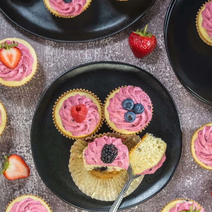 Overhead view of assorted mixed berry cupcakes topped with pink berry buttercream and fresh fruit, arranged on dark plates against a textured background