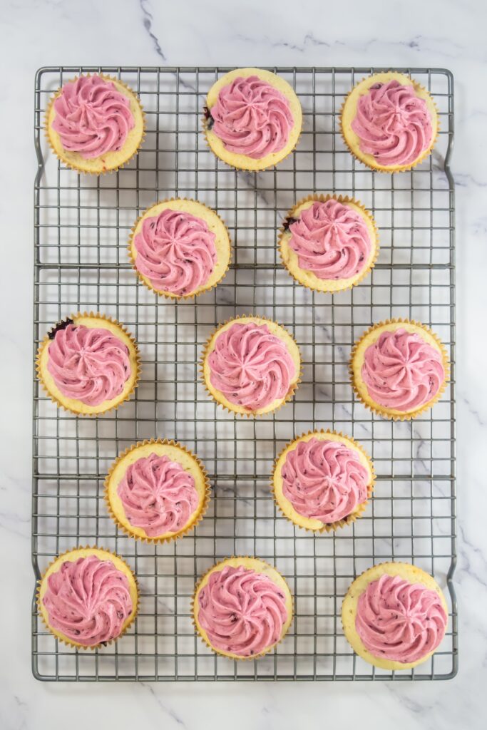 A close-up angled shot of berry cupcakes with pink swirled frosting and fruit toppings, displayed on a wire rack