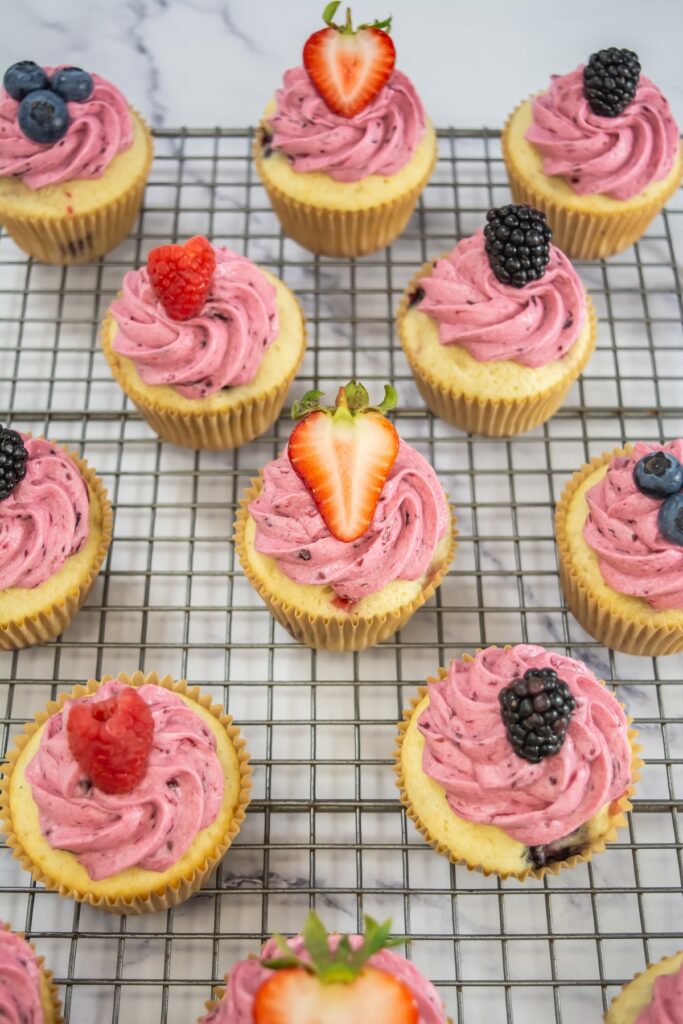 A halved cupcake with visible berry pieces sits on an unwrapped liner, surrounded by whole frosted cupcakes on black plates