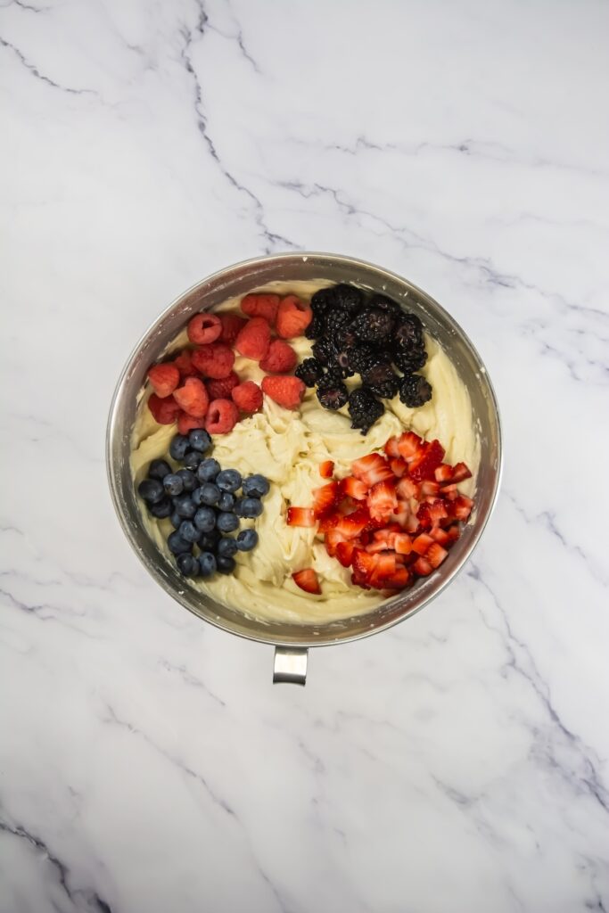 A colorful mix of fresh berries — blueberries, raspberries, blackberries, and chopped strawberries — placed on top of smooth cupcake batter in a metal bowl