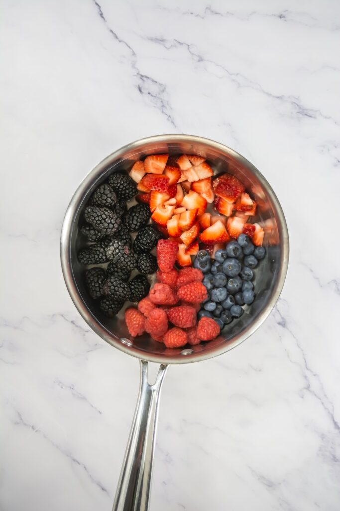 A saucepan filled with strawberries, blackberries, raspberries, and blueberries, set over a marble counter for making a fresh berry compote or filling
