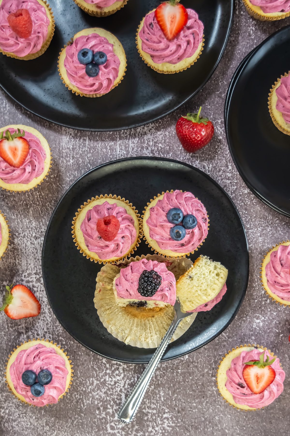 Overhead view of assorted mixed berry cupcakes topped with pink berry buttercream and fresh fruit, arranged on dark plates against a textured background