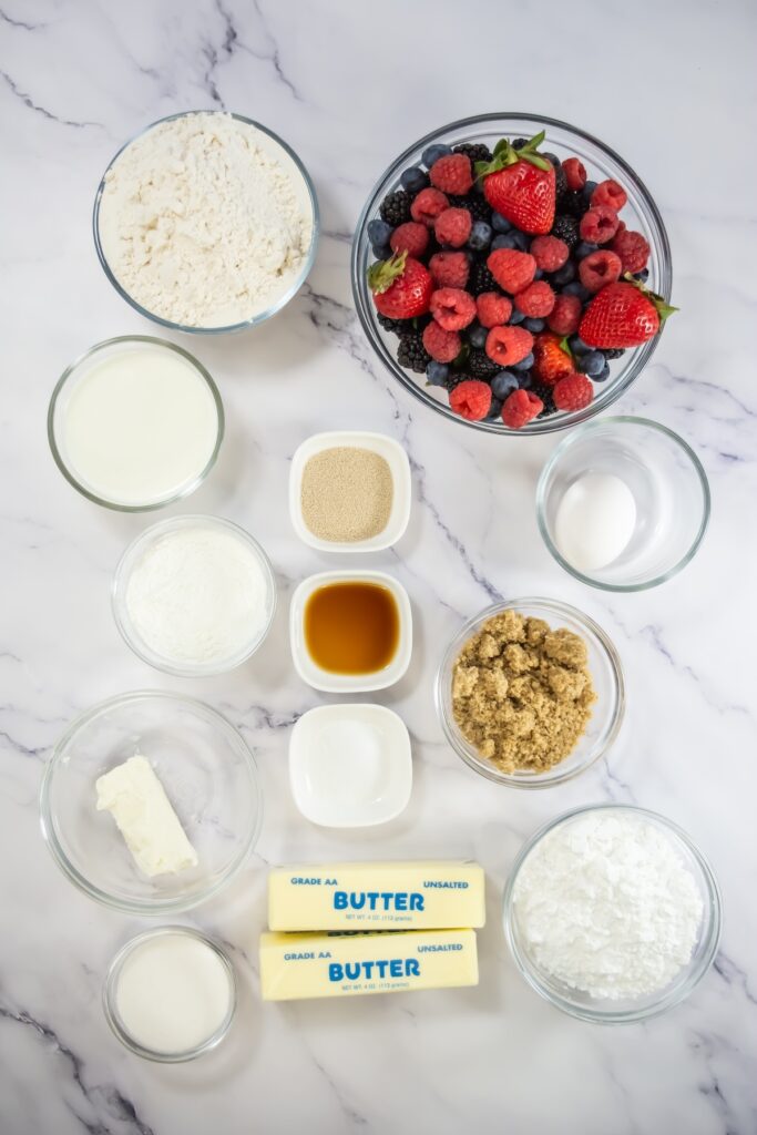 Overhead view of ingredients for mixed berry sweet rolls on a marble surface, including fresh berries, flour, butter, brown sugar, yeast, cream cheese, vanilla extract, and powdered sugar