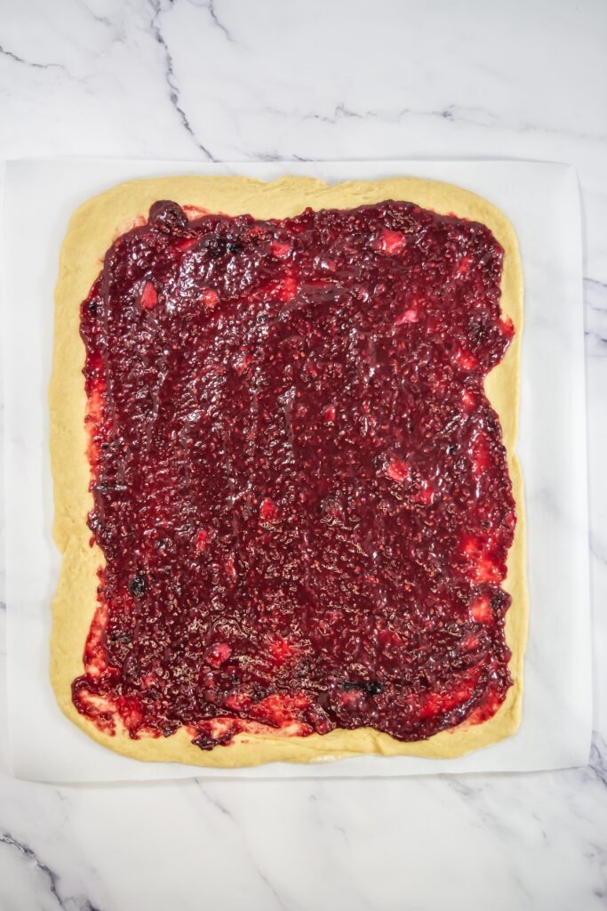 Rolled-out sweet roll dough topped with a thick layer of chunky mixed berry filling spread evenly to the edges on parchment paper