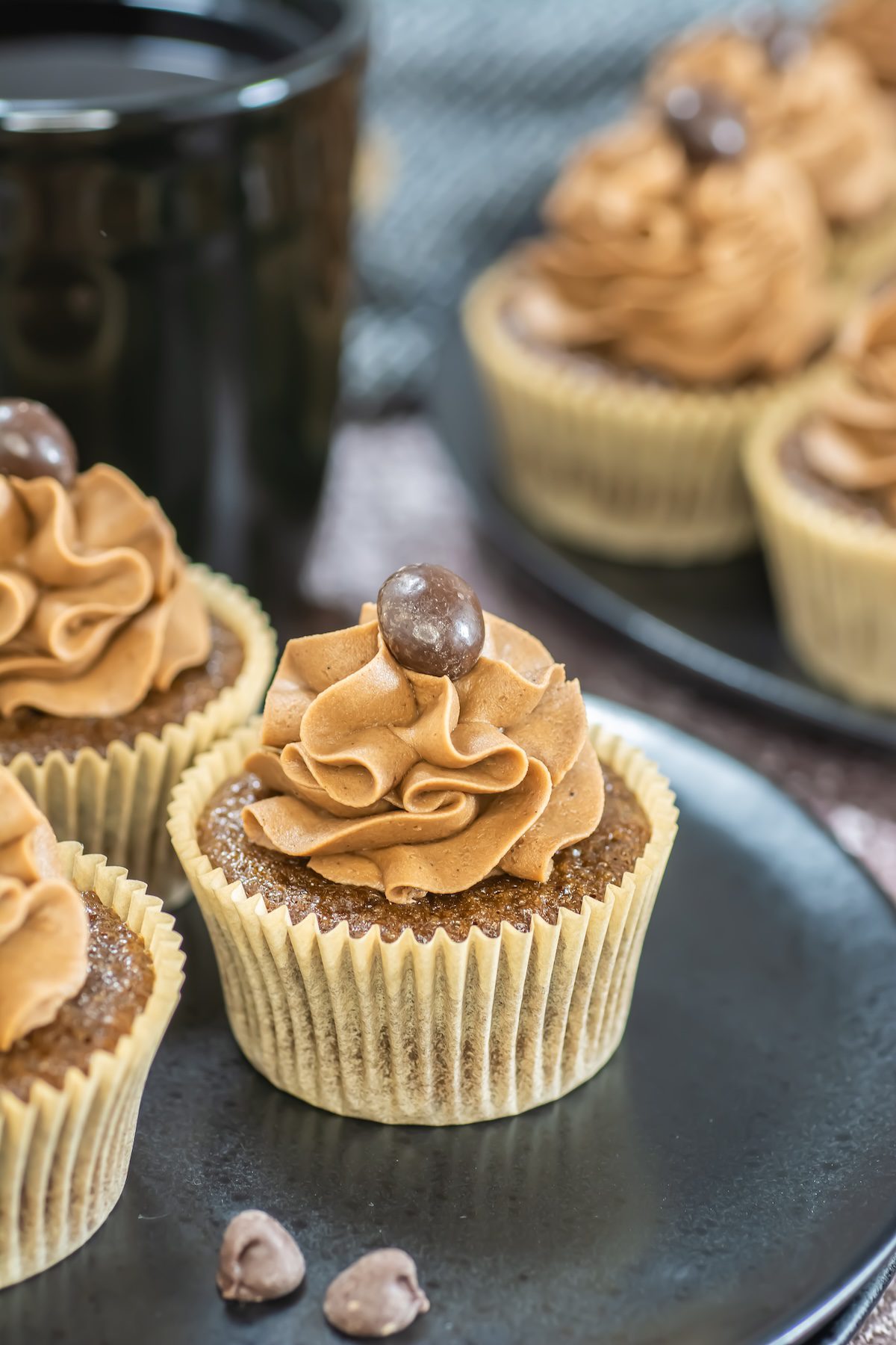 A display of mocha cupcakes topped with swirled coffee frosting and chocolate espresso beans, with one cut cupcake in the foreground and a bowl of coffee beans nearby