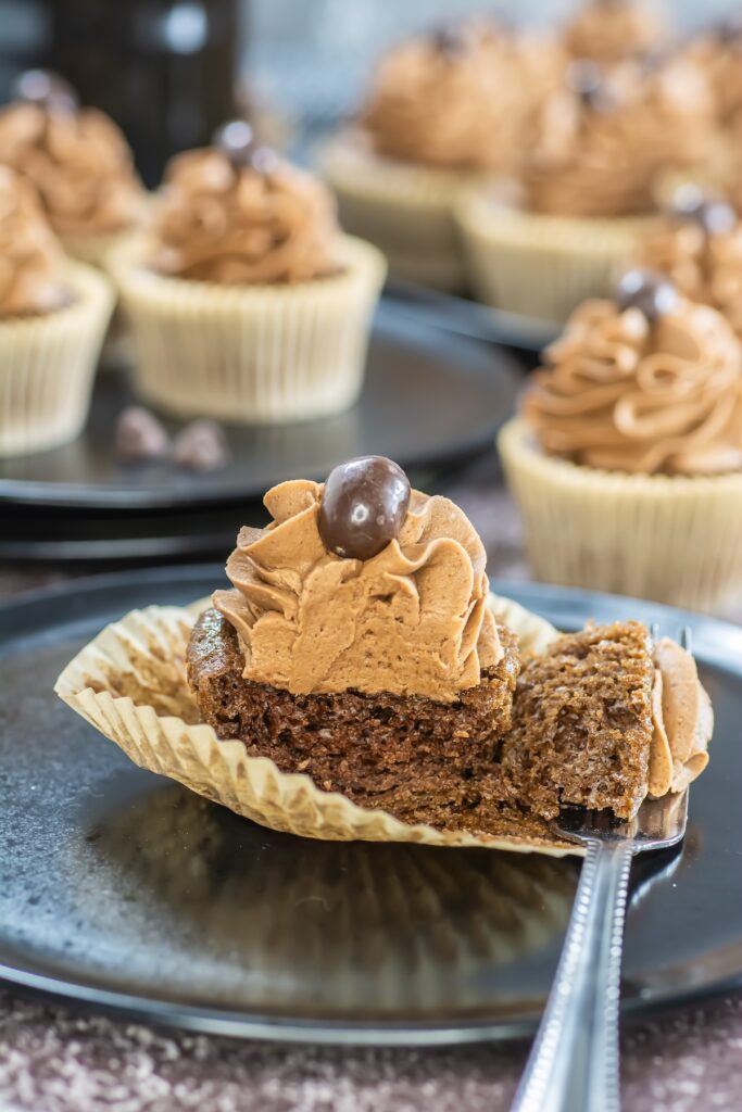 A bitten mocha cupcake showing its rich, fluffy texture, with coffee frosting and a chocolate-covered coffee bean on top, set on a dark plate with a fork