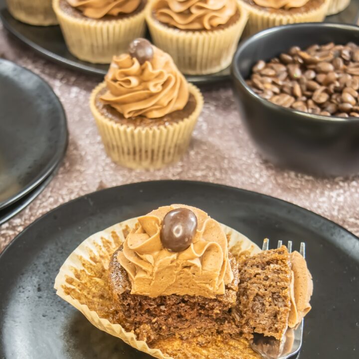 A close-up of moist mocha cupcakes topped with silky coffee buttercream swirls and a chocolate-covered espresso bean, served on a dark plate with scattered chocolate chips