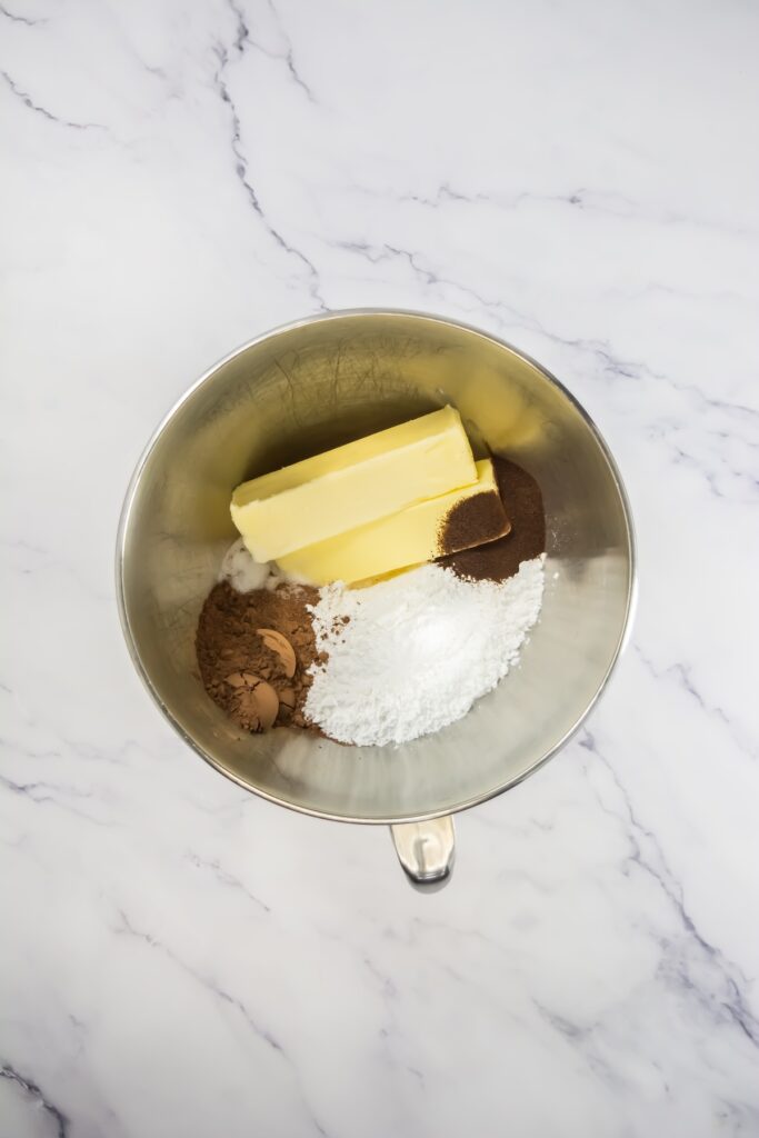 Mixing bowl with ingredients for coffee frosting, including butter, cocoa powder, espresso powder, and powdered sugar
