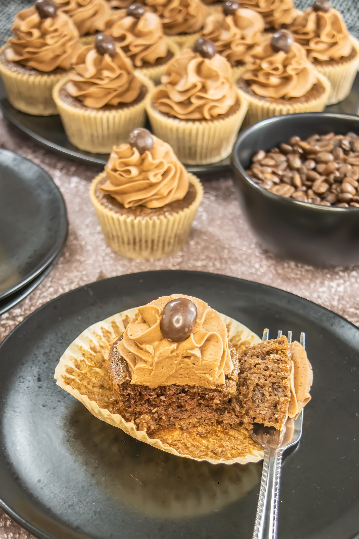 A close-up of moist mocha cupcakes topped with silky coffee buttercream swirls and a chocolate-covered espresso bean, served on a dark plate with scattered chocolate chips